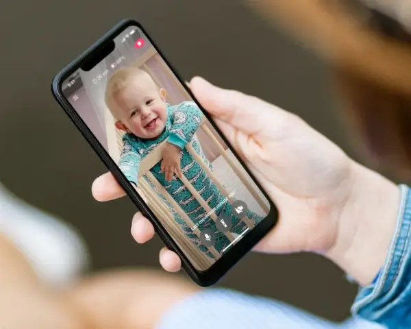 Ein lächelndes Baby im Strampler schaut aus dem Gitterbett. Eine Hand hält das Handy als Babyphone, um das Kind im Blick zu behalten.