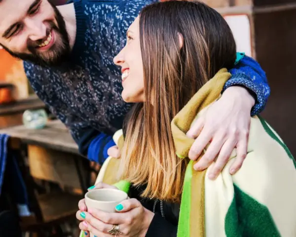 Pareja sonriente compartiendo una manta y una taza de té. Él le abraza por detrás. Ella le dice "muchas gracias en francés".