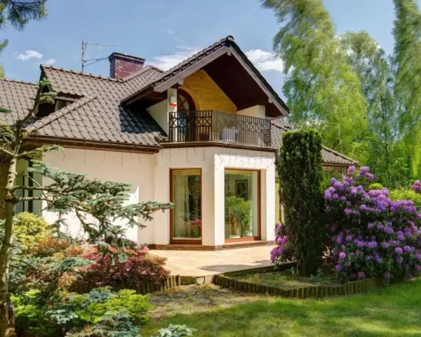 Dom z balkonem i tarasem, otoczony bujną zielenią i kwitnącymi rododendronami. Występ ściany podkreśla nowoczesny charakter budynku.