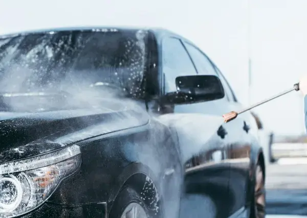 Man med t-grön tröja tvättar en svart bil med högtryckstvätt. Vatten och skum sprutar över bilen.