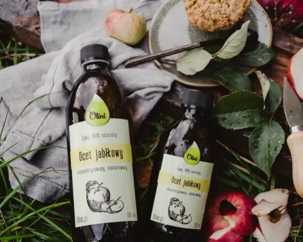 Dwie butelki naturalnego octu jabłkowego Olini, idealnego dla dzieci, leżą wśród jabłek i liści.
