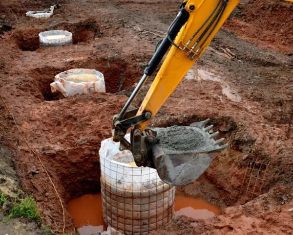 Koparka zasypuje betonem fundament palowy w wykopie. W tle widać kolejne przygotowane do zalania pale.