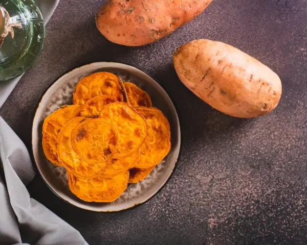 Chrupiące chipsy z batatów na talerzu, obok surowych bulw. Niska zawartość cukru sprawia, że bataty indeks glikemiczny mają korzystny wpływ na dietę.