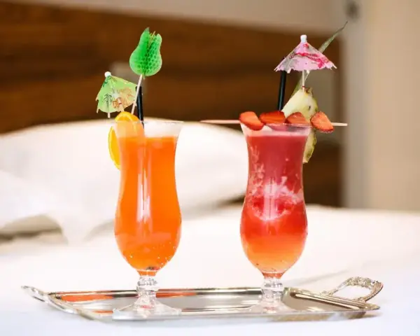 Dwa orzeźwiające drinki: jeden pomarańczowy z pomarańczą, drugi czerwony z truskawkami i ananasem, oba z parasolkami. Idealne na wakacje, może z dodatkiem triple sec.