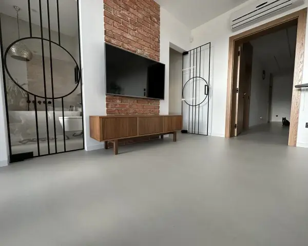 Nowoczesne wnętrze z betonową podłogą, co to jest mikrocement, która nadaje mu surowego charakteru. W tle widać łazienkę i salon.