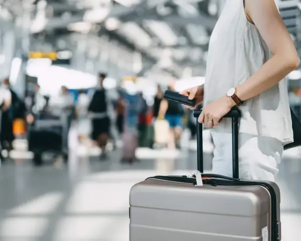 A traveler holds a passport and suitcase, mindful of carry-on size limits in a busy airport.
