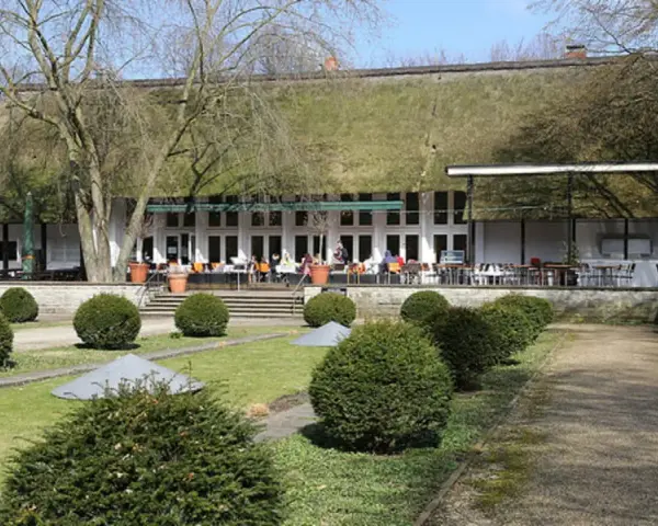 Das Teehaus im Englischen Garten Berlin mit seinem Reetdach und der Terrasse, auf der Gäste sitzen, ist von gepflegten Rasenflächen und Bäumen umgeben.