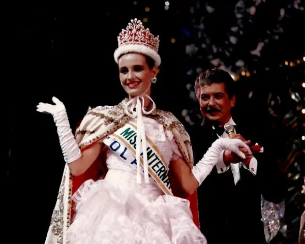 Uśmiechnięta Miss International w koronie i białej sukni, z szarfą "Miss Polonia". Obok niej mężczyzna w smokingu.