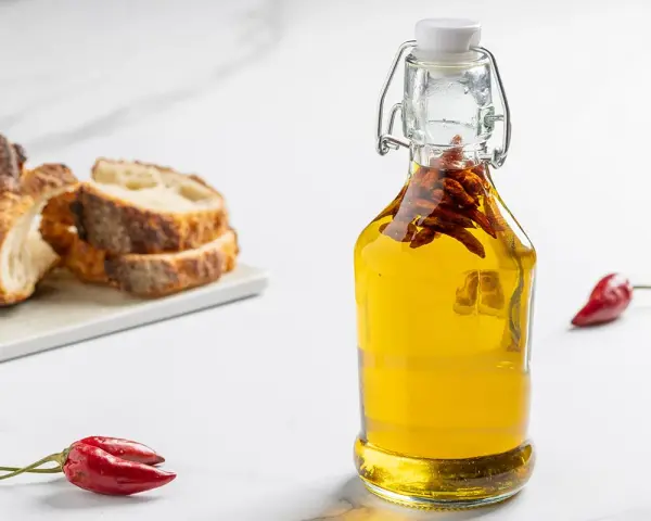 Olio al peperoncino in bottiglia, con peperoncini rossi e pane rustico a fette sullo sfondo.