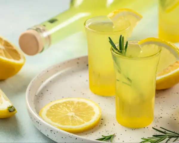 Orzeźwiający limoncello co to? Włoski likier cytrynowy w kieliszkach, ozdobiony cytryną i rozmarynem, obok butelki i świeżych cytryn.