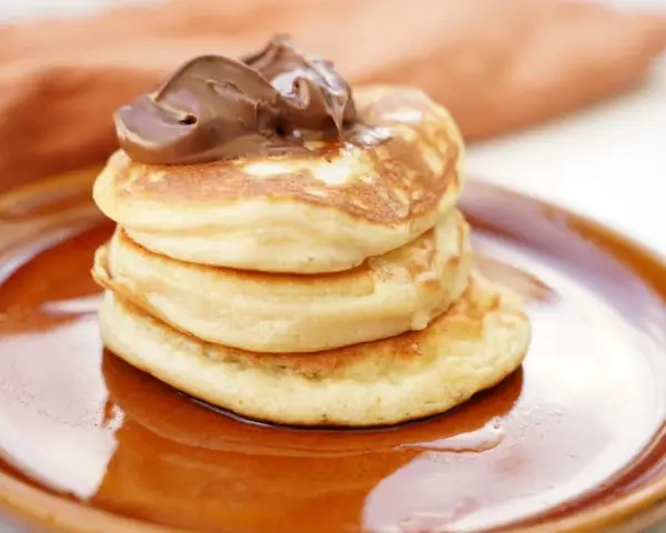 Stack of three fluffy American pancakes topped with chocolate spread on a brown plate.