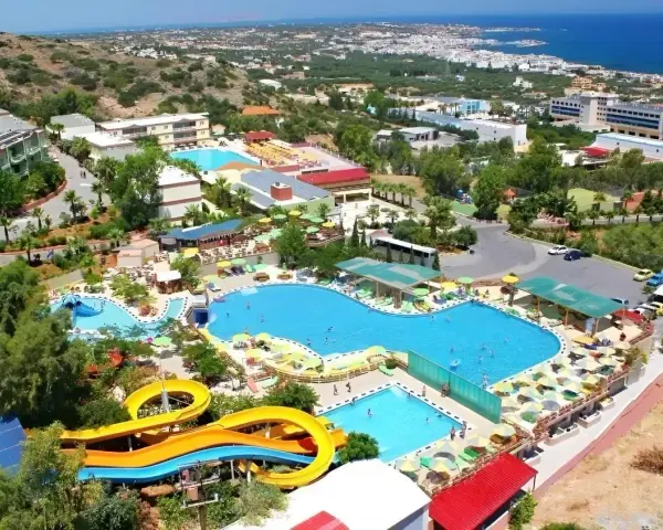 Widok z lotu ptaka na kompleks basenów z atrakcjami wodnymi i leżakami. Wiele osób korzysta z uroków słońca i wody. W tle widać budynki hotelowe i morze. Aqua Sun Village opinie potwierdzają, że to idealne miejsce na wakacje.