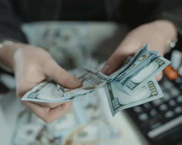 Dłonie liczą banknoty 100-dolarowe. Obok leży kalkulator, sugerując obliczanie kwoty, być może jako kara pieniężna za wyrządzoną krzywdę.