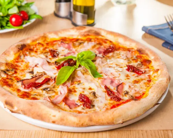 Pizza garnie de jambon, tomates séchées et champignons, parfaite pour une recette pâte à pizza rapide. Une salade fraîche est servie à côté.