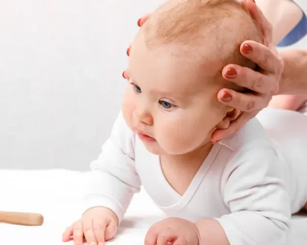 Dłoń delikatnie dotyka główki niemowlęcia, sprawdzając ciemiączko tylne. Obok leży drewniana zabawka.
