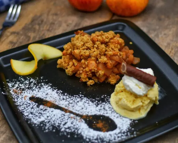 Ein köstlicher Apfel Crumble mit Haferflocken, serviert mit Vanilleeis, Sahne und einem Schokostäbchen.