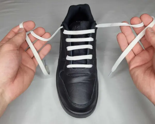 Mani che mostrano come mettere i lacci alle scarpe senza fiocco, con un laccio bianco che attraversa gli occhielli di una scarpa nera.