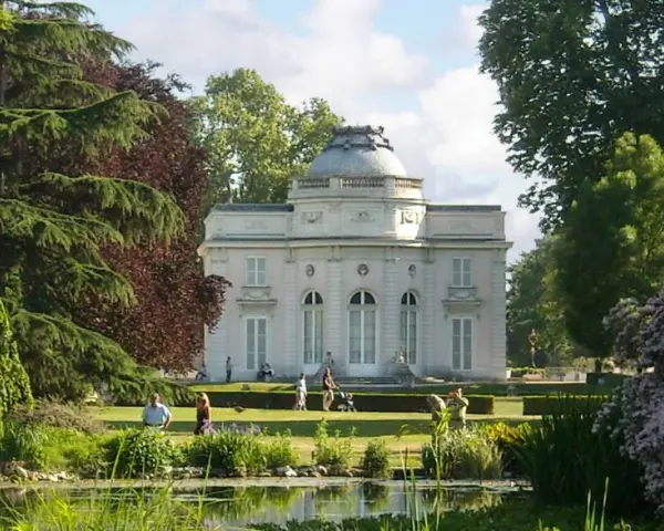 Un elegante edificio blanco con cúpula se alza en un exuberante jardin des plantes, con un estanque en primer plano y gente paseando.