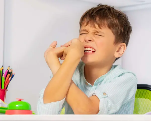 Junge mit zusammengepressten Augen und Händen vor dem Gesicht, ein Zeichen dafür, wie mit Tics bei Kindern umgehen. Buntstifte und eine Klingel liegen auf dem Tisch.