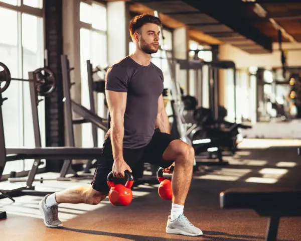 Mężczyzna wykonuje wykroki z kettlebellami, realizując swój plan treningowy na siłownię.