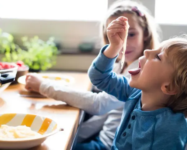 Zwei Kinder am Tisch, die schnell Nudeln essen. Der Junge nimmt eine Gabel voll Spaghetti zum Mund.