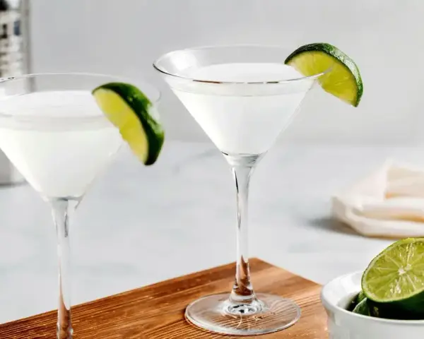 Dwa kieliszki z białym drinkiem, ozdobione plasterkami limonki. Obok miseczka z limonkami i shaker. Idealny kamikaze shot na gorący wieczór.