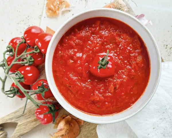 Ein Teller mit leuchtend rotem Sugo di Pomodoro, garniert mit einer einzelnen Kirschtomate. Daneben ein Bund frischer Tomaten und Zwiebeln.