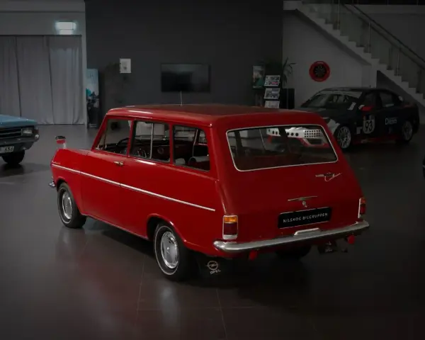 Röd veteranbil med ålder, en Opel Caravan, står i en bilhall bredvid en blå bil och en tävlingsbil.