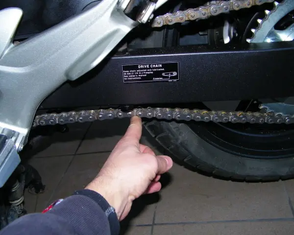 Sprawdzanie luzu łańcucha w motocyklu. Palec pokazuje, jak sprawdzić, czy łańcuch jest prawidłowo naciągnięty.