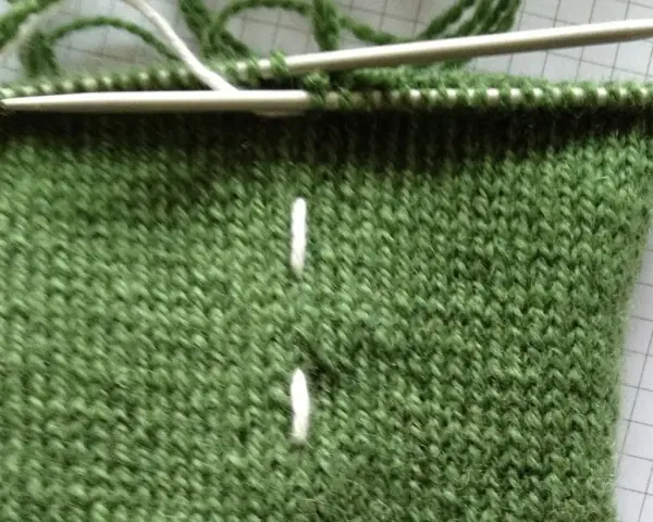 Grönt stickarbete med två vita markeringar, stickor och z tvinnat garn.