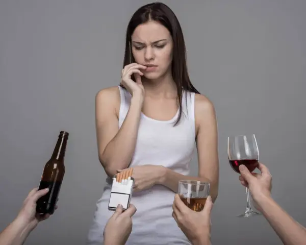 Kobieta w białej koszulce zastanawia się, jak przestać pić, gdy wokół niej wyciągane są papierosy, piwo i wino.