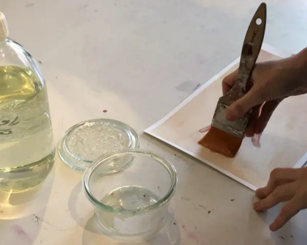 Application d'une finition acrylique sur une œuvre. Une main tient un pinceau large, prêt à recouvrir le papier d'une couche protectrice.
