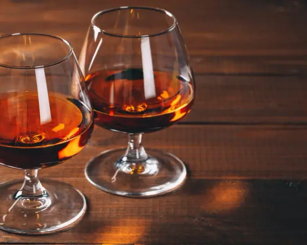 Dwa kieliszki z bursztynowym płynem, przypominającym bourbon lub whiskey, stoją na drewnianym stole.