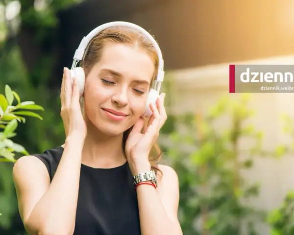 Kobieta w słuchawkach na uszach, z zamkniętymi oczami, cieszy się chwilą słuchania muzyki w otoczeniu zieleni.