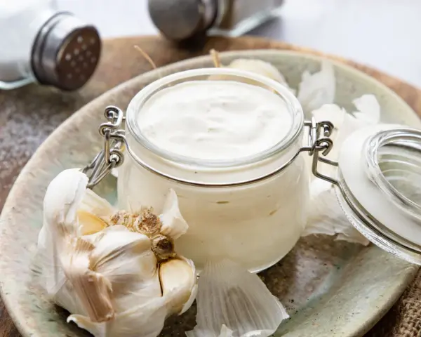 Frische Knoblauchsauce selber machen: cremige Sauce im Glas mit Knoblauchzehen und Salzstreuer im Hintergrund.