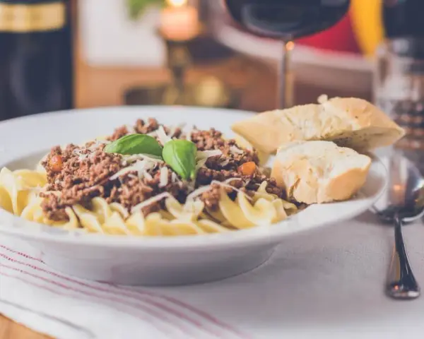 Pyszny makaron z sosem bolognese, posypany parmezanem i udekorowany bazylią. Czy to spaghetti, czy tagliatelle? Obok kromki chleba i kieliszek czerwonego wina.