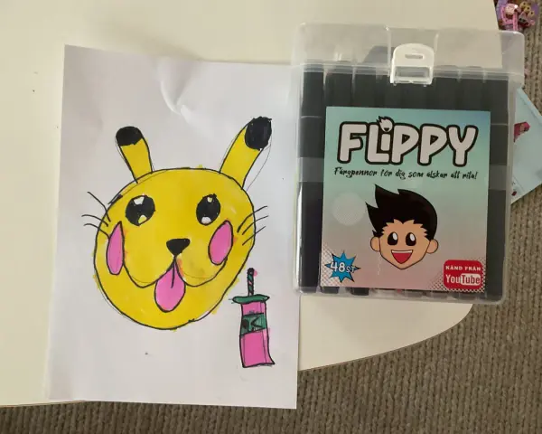 En gul Pikachu-liknande figur med rosa kinder och en tunga som sticker ut, bredvid en låda med färgpennor "FLIPPY" för dig som älskar att rita.