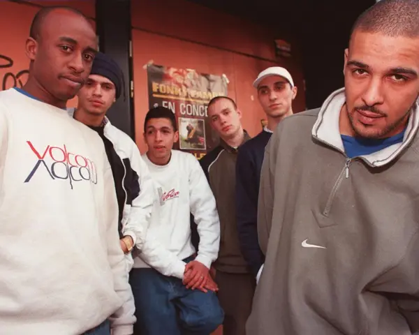 Un groupe de rap français des années 90/2000 pose devant un mur tagué. Ils portent des sweats et des jeans, typiques de l'époque.