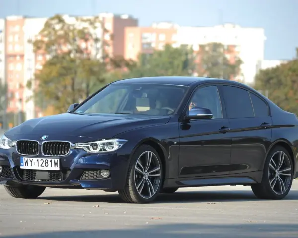 Ciemnoniebieskie BMW 318d z polskimi tablicami rejestracyjnymi stoi na drodze. W tle widać budynki i drzewa.