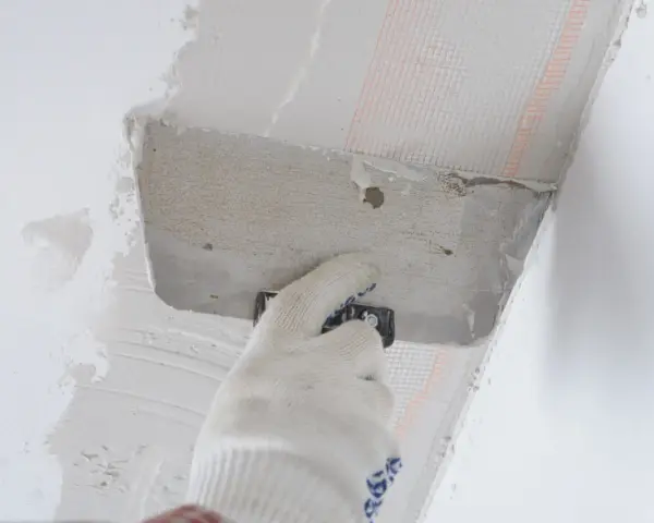 Ręka w rękawicy nakłada zaprawę na styropian przy suficie. Trwa klejenie styropianu do sufitu na piankę.