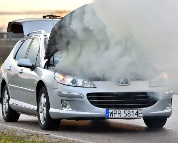Srebrny samochód Peugeot z otwartą maską, z której wydobywa się gęsty dym.