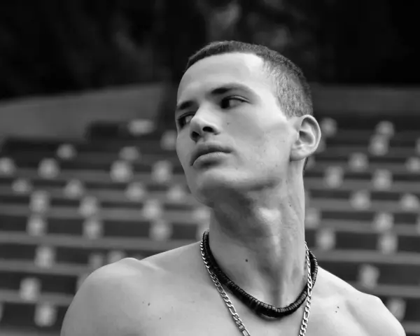 Joven con un corte de pelo buzz cut largo, mirando hacia un lado con una expresión pensativa. Lleva un collar de cadena.