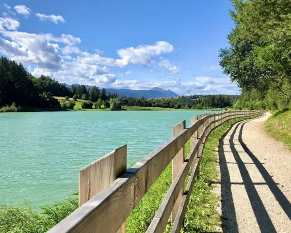 Sentiero sterrato con staccionata in legno lungo il lago di Tavon, circondato da alberi e montagne. Cielo azzurro con nuvole bianche.
