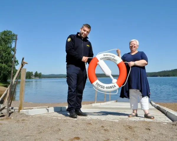 Två personer håller i en räddningsboj vid en sjö. En man i uniform och en kvinna ler. Trygg-Hansa vid Nörybyns camping.