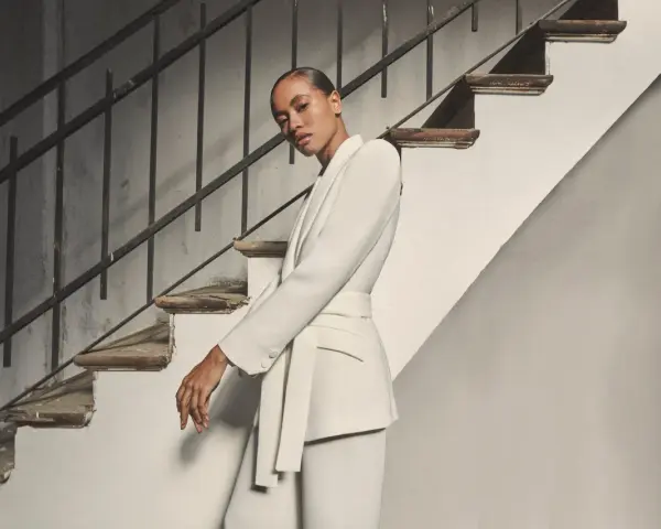 Mujer posando con un elegante outfit blazer blanca, pantalón a juego y cinturón, junto a una escalera antigua.