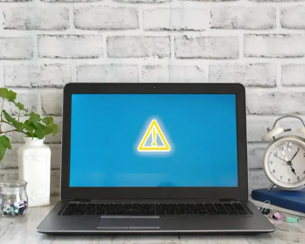 Laptop z symbolem ostrzeżenia na ekranie, obok budzik i rośliny.