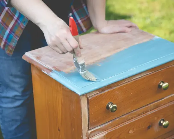 Persona pinta con brocha azul una mesita de noche de madera, transformándola en una de las mesitas de noche pintadas a la tiza.