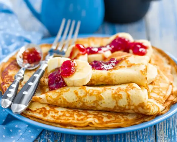 Stos idealnie przygotowanego ciasta na naleśniki, polanego dżemem i kawałkami banana. Obok leżą sztućce.