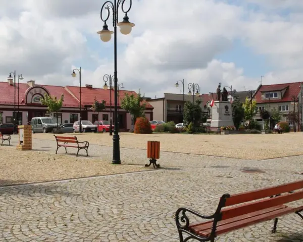 Rynek w Ślesinie z pomnikiem, ławkami i budynkami. Atrakcje tego miejsca zachęcają do odpoczynku.