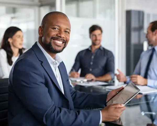 The executive board chair smiles, holding a tablet in a meeting with colleagues.